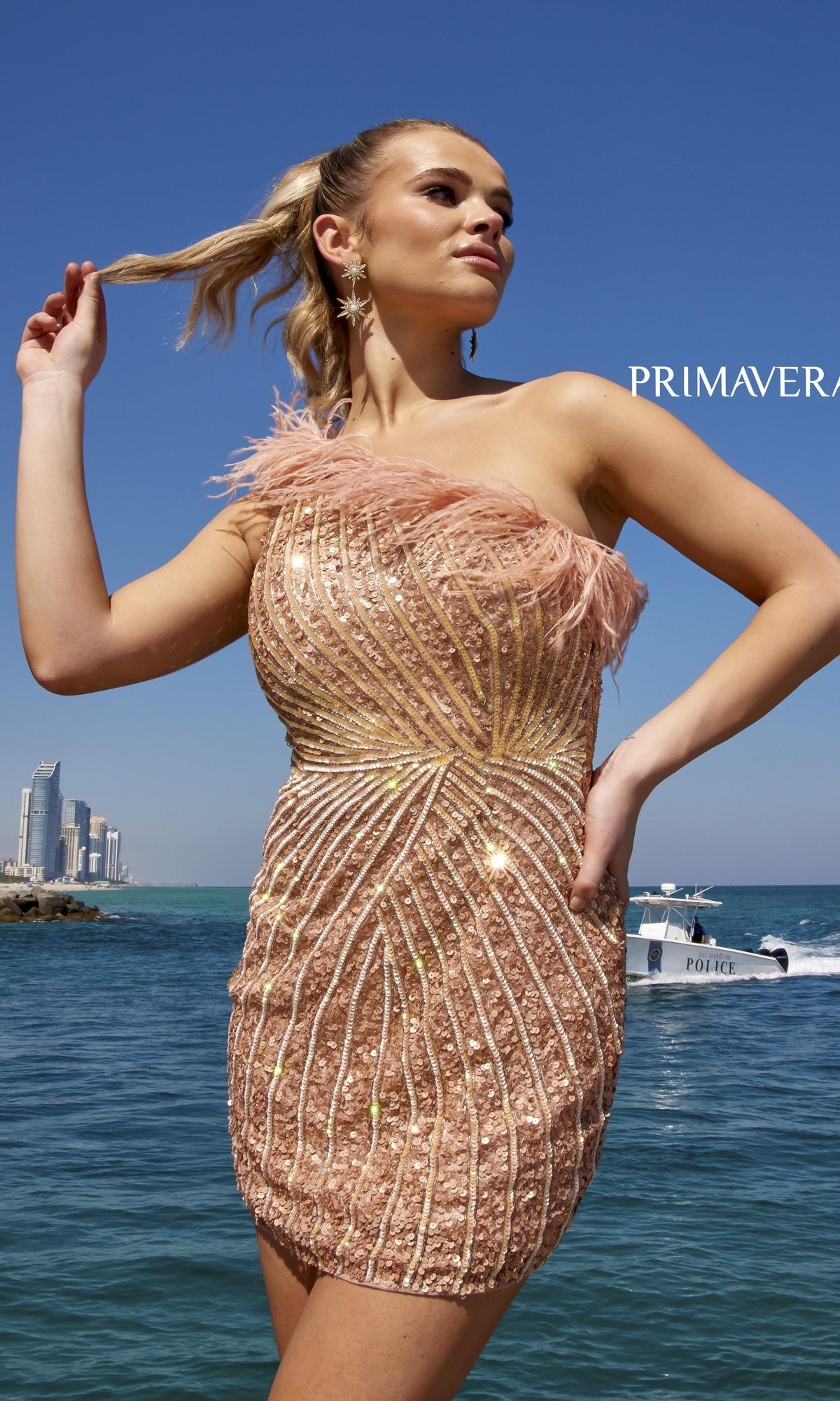 A woman in the One-Shoulder Sequin Party Dress with Feathers 4002 poses by the water with a city skyline and police boat in the background under a clear blue sky.